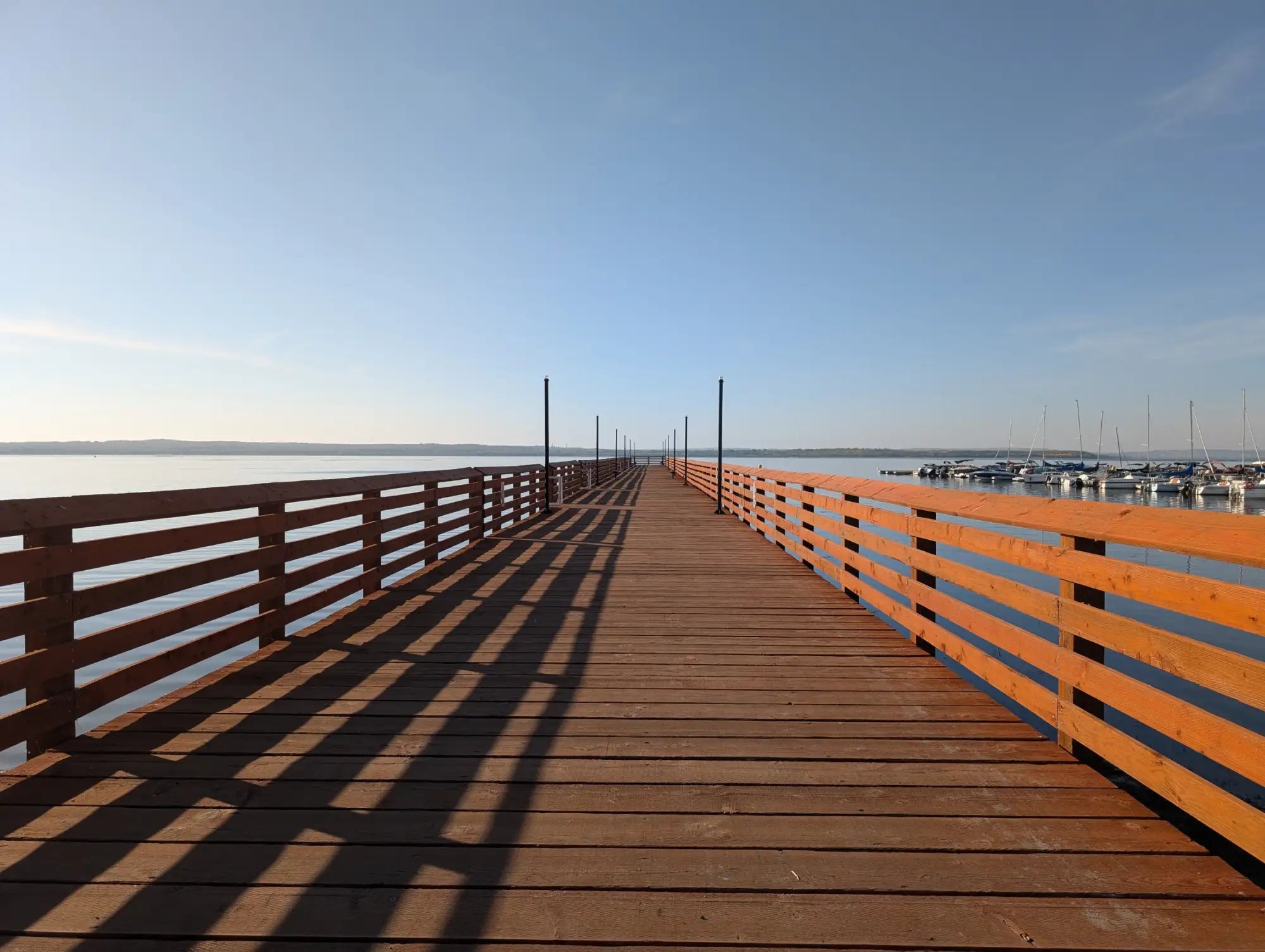 A long wooden pier with warm brown planks and matching railings stretches out over calm water under a clear blue sky.