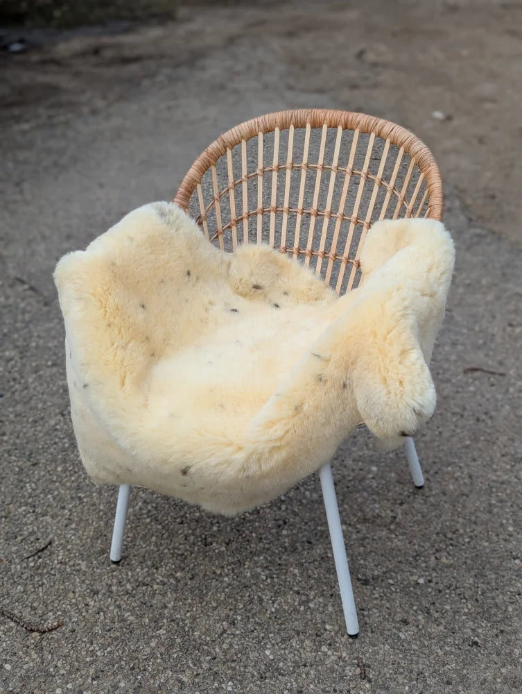 A cozy cream-colored naturally spotted medical sheepskin draped over a woven wicker chair with white metal legs