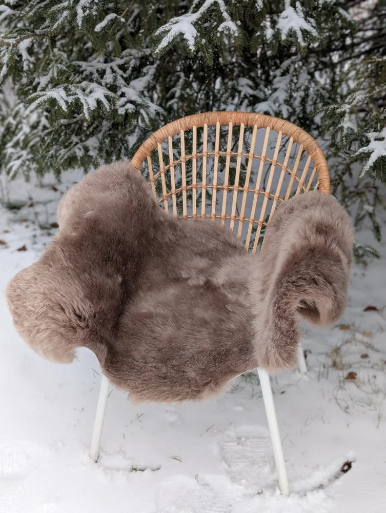 A cozy medium taupe merino sheepskin chair cover draped over a wicker chair in snowy outdoor setting