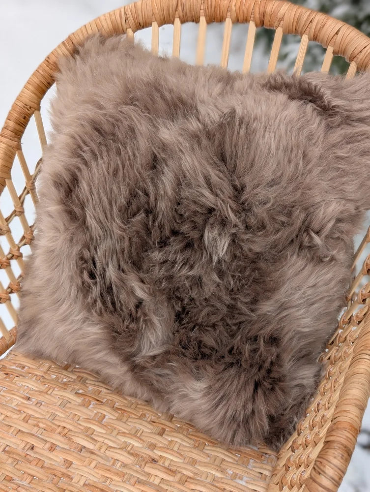 A medium taupe merino sheepskin cushion with soft fluffy texture and cotton backs on a woven wicker chair