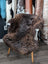 A plush dark brown british isles sheepskin rug draped over a white chair with wooden legs arriving soon