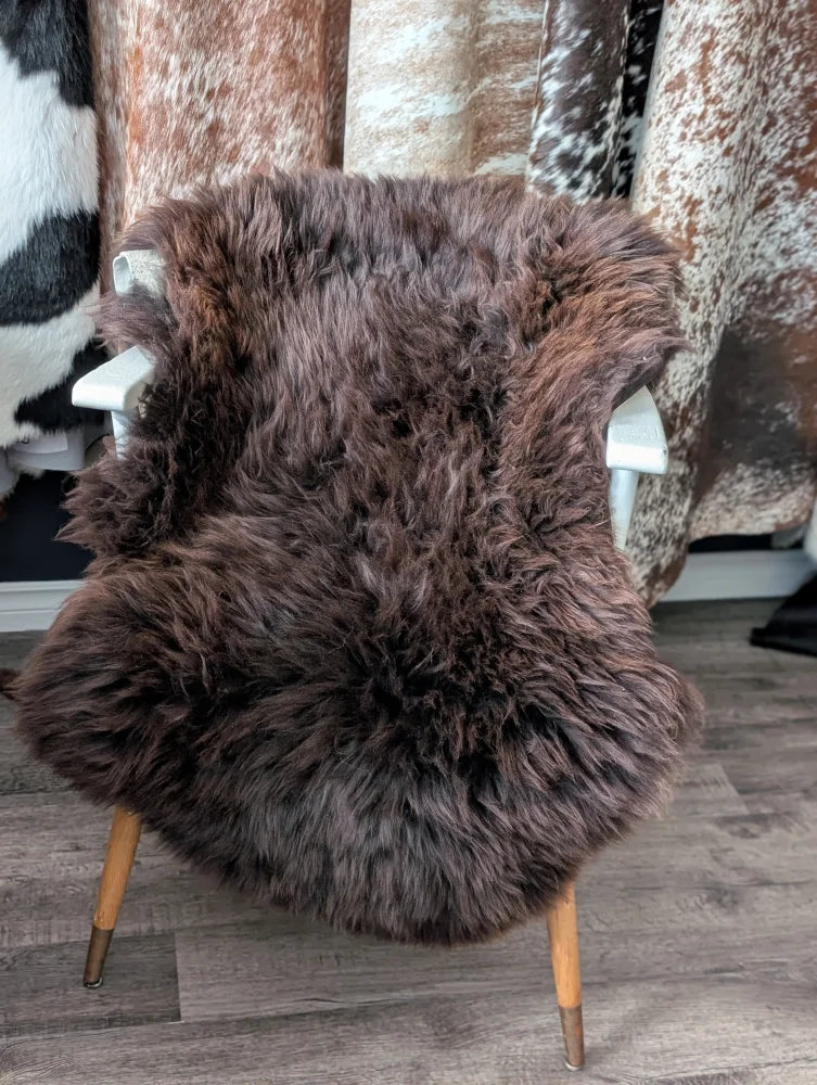 A plush dark brown british isles sheepskin rug with thick fluffy texture draped over the back of a white chair