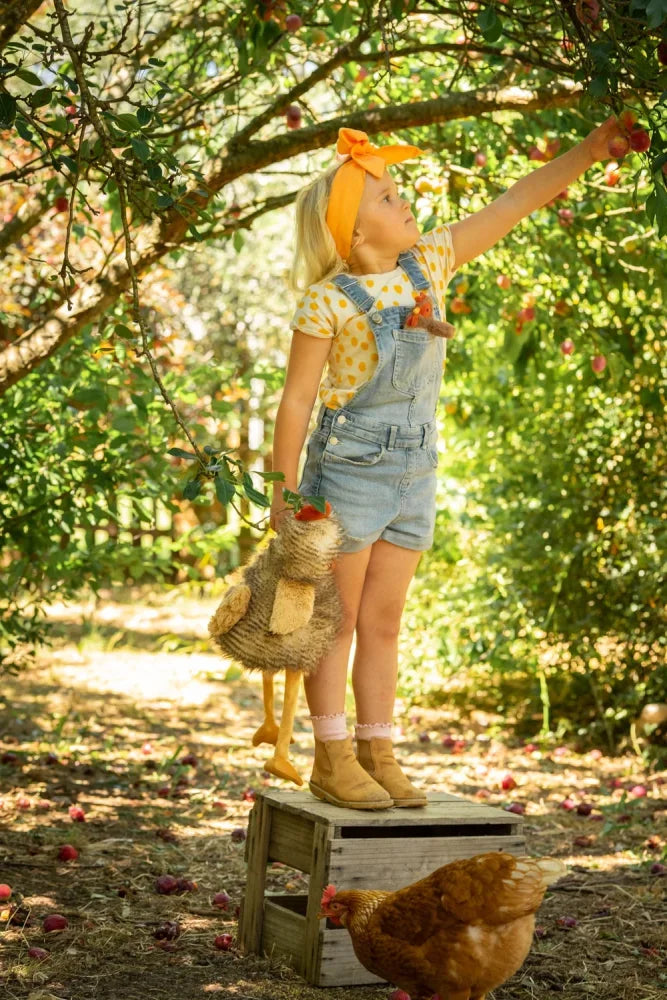 A young girl in denim overalls and a yellow polka-dot shirt reaches for apples on a crate in rupert the rooster