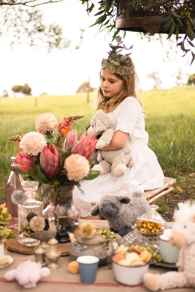 A young girl in a white dress holds Sammy the Sheep plush toy outdoors wearing a leafy headband