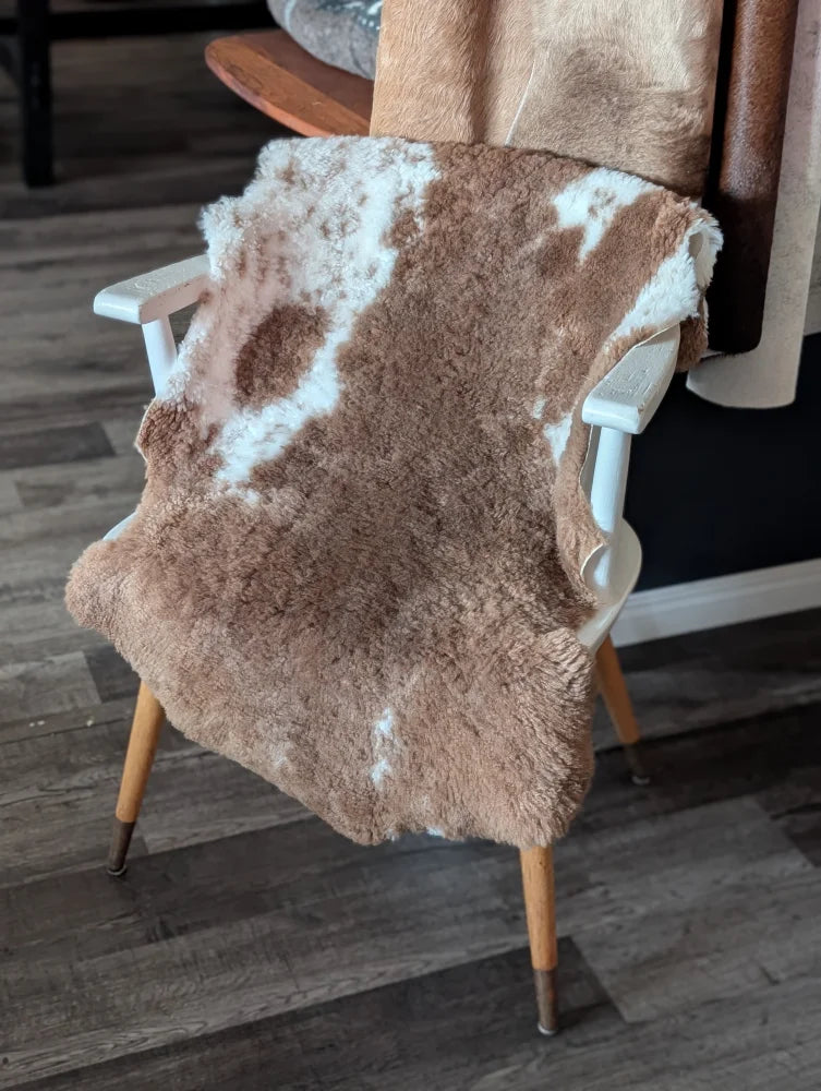 A plush brown and white tibet highland spotted sheepskin rug draped over a white wooden chair with light wood legs