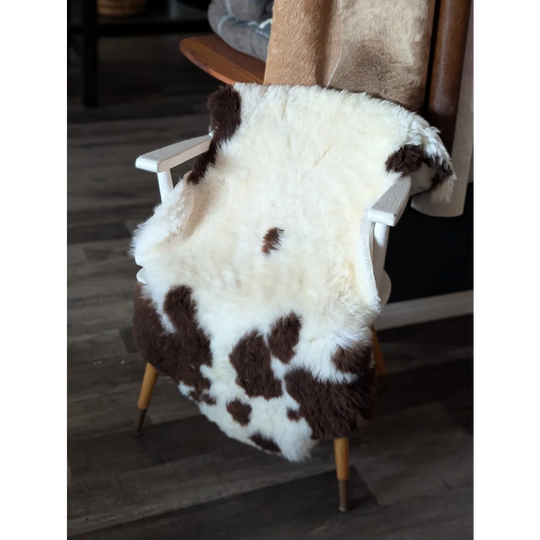 A plush white and brown tibet highland spotted sheepskin rug draped over a white wooden chair with light wood legs