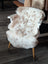 A plush cream-colored tibet highland spotted sheepskin rug draped over a white wooden chair with light wood legs