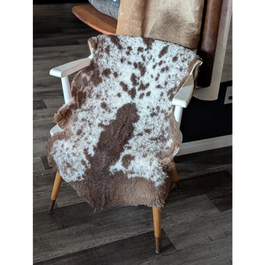 A plush brown and white tibet highland spotted sheepskin rug draped over a white wooden chair with light wood legs