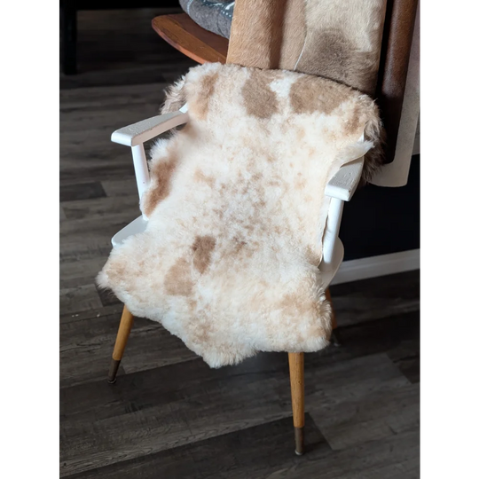 A cream and tan tibet highland spotted sheepskin rug draped over a white wooden chair with light wood legs