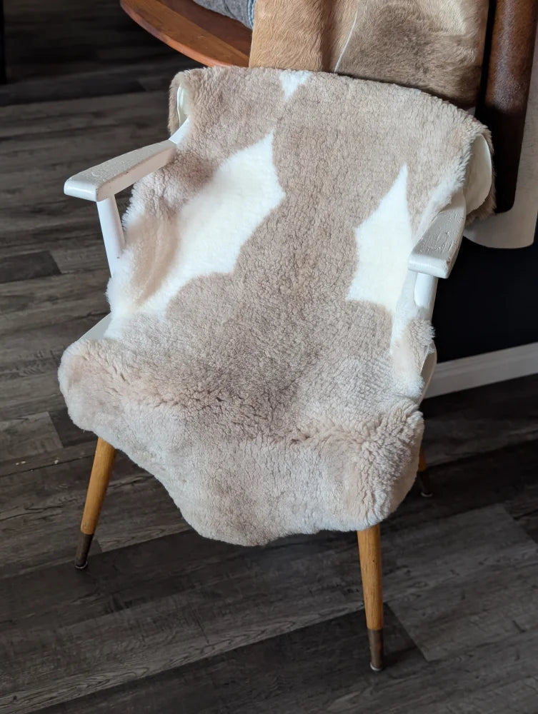 A cozy cream and white tibet highland spotted sheepskin blanket draped over a white chair with light wood legs