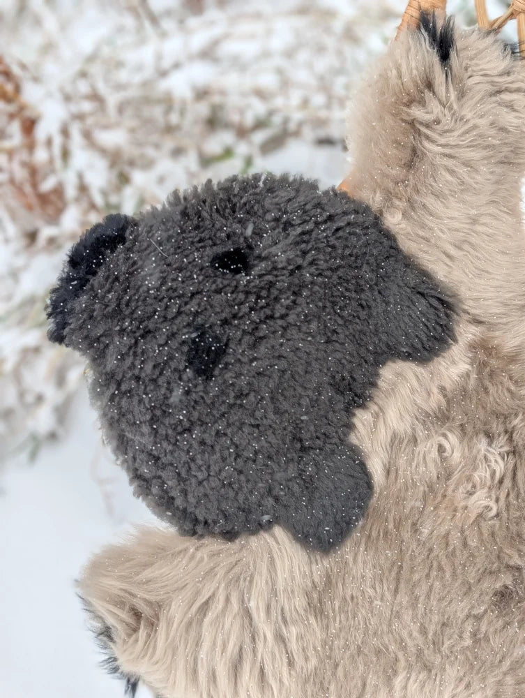 A hand made dark gray plush teddy bear head on a light beige merino sheepskin grizzly bear rug surface