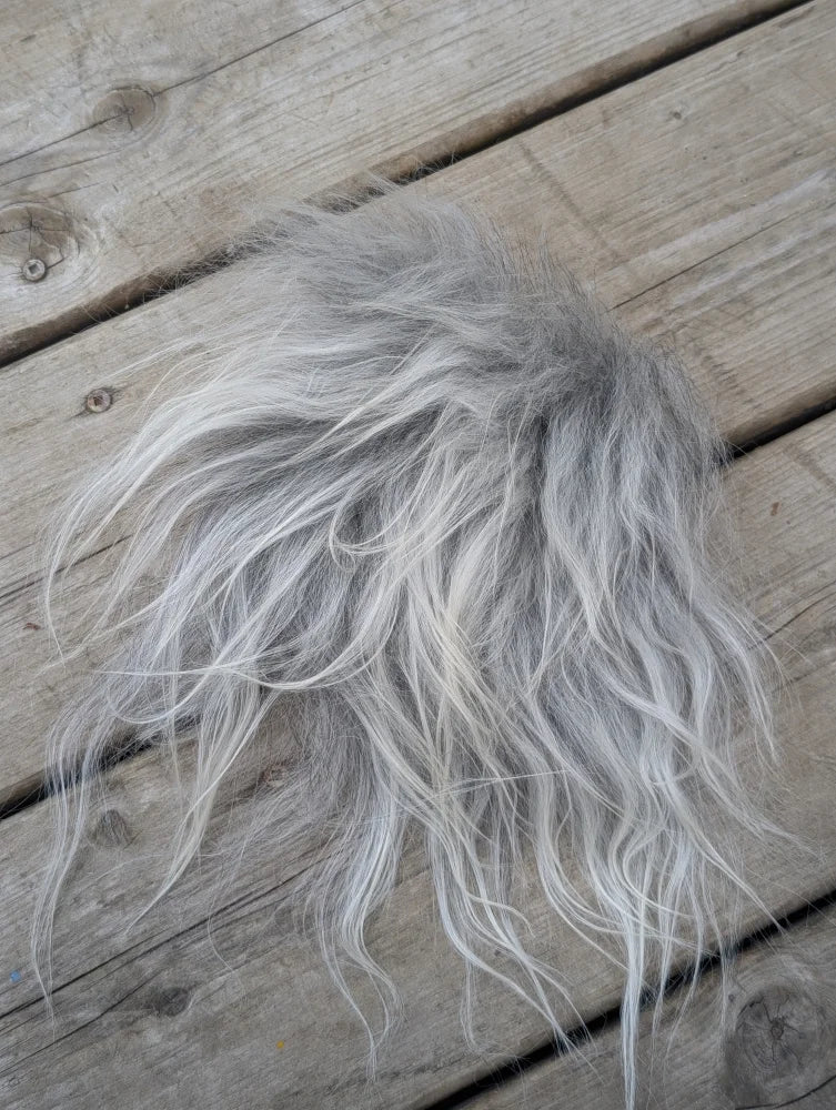 A fluffy silver-gray sheepskin sample square with long wavy faux fur strands lying on a wooden surface