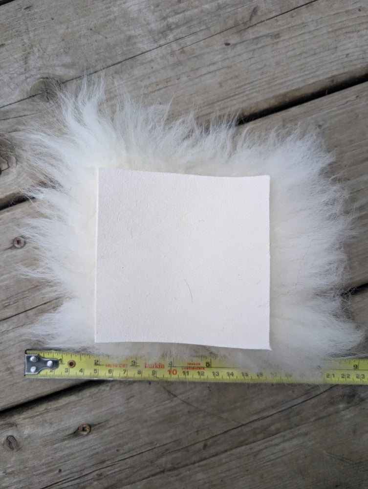 White rectangular sheepskin sample square with fluffy fur edges resting on wood with measuring tape below