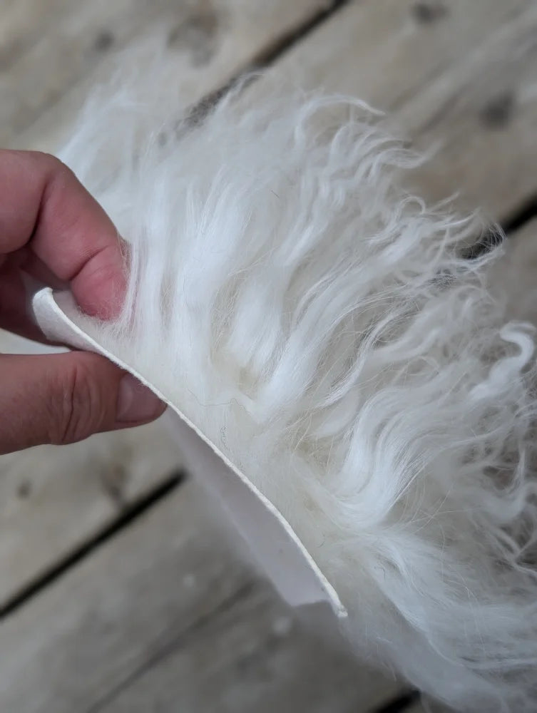 A hand holds a white fluffy curly sheepskin sample square made of soft wool fiber material
