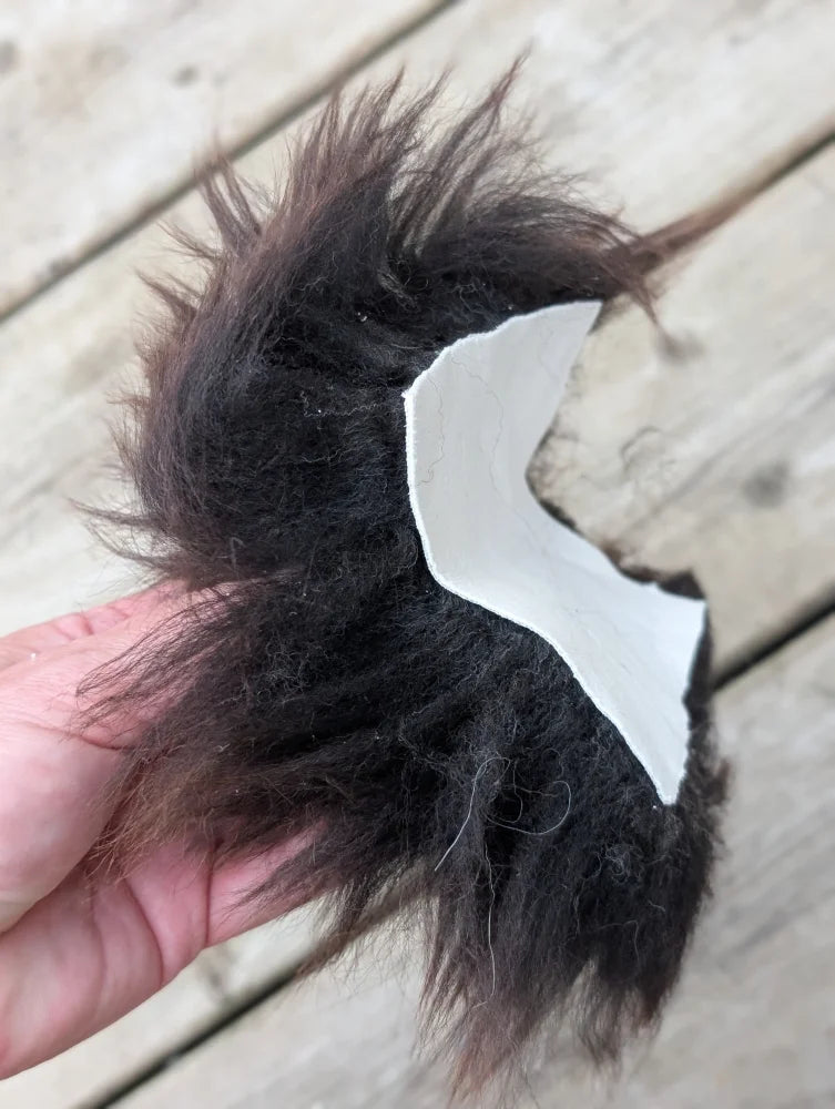 A hand holds a dark brown fluffy faux fur sheepskin sample square with a white paper cutout attached