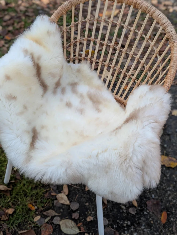 A cream british isles sheepskin rug with brown speckles resting on a woven wicker chair softbrit 47x28 inches