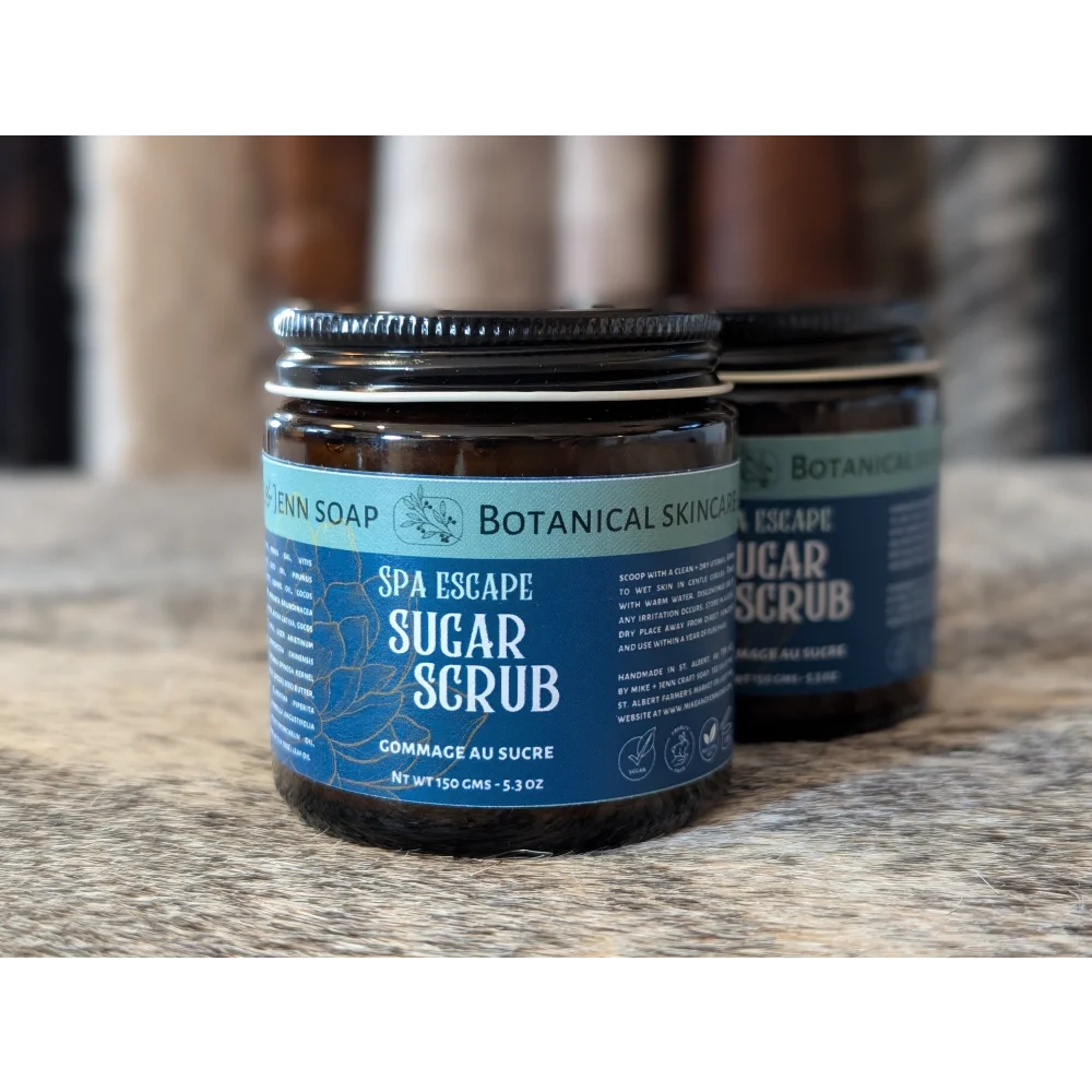 A dark amber glass jar with a silver lid and blue label containing spa escape sugar scrub with dead sea salt and essential oil blend