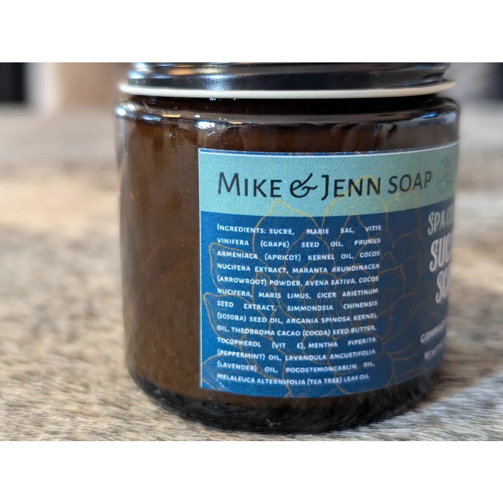 A brown glass jar with black lid and blue label mike &amp; jenn soap contains dark textured spa escape sugar scrub with dead sea salt