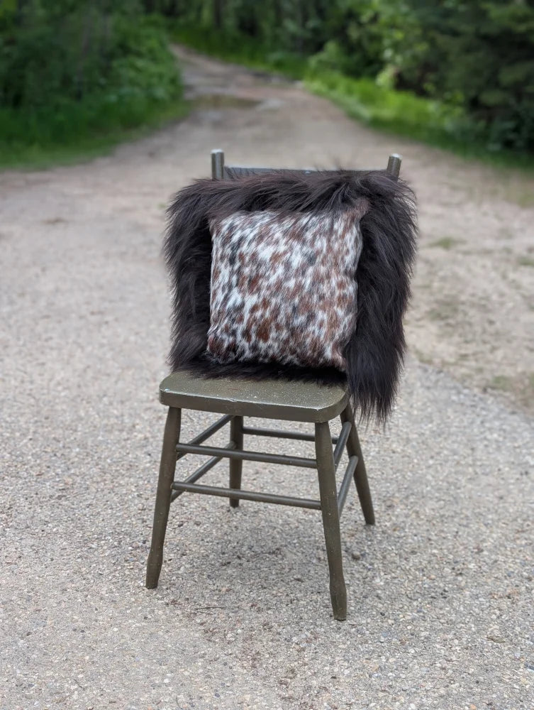 A cozy speckled cowhide cushion with white and brown spots sits on dark fur throw on olive green chair with long wool accents