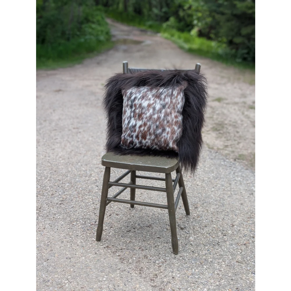 A cozy speckled cowhide cushion with white and brown spots sits on dark fur throw on olive green chair with long wool accents