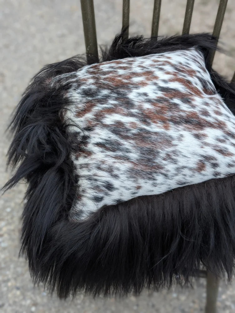 A plush square cushion in natural cowhide with white and brown speckles and long wool black icelandic trim
