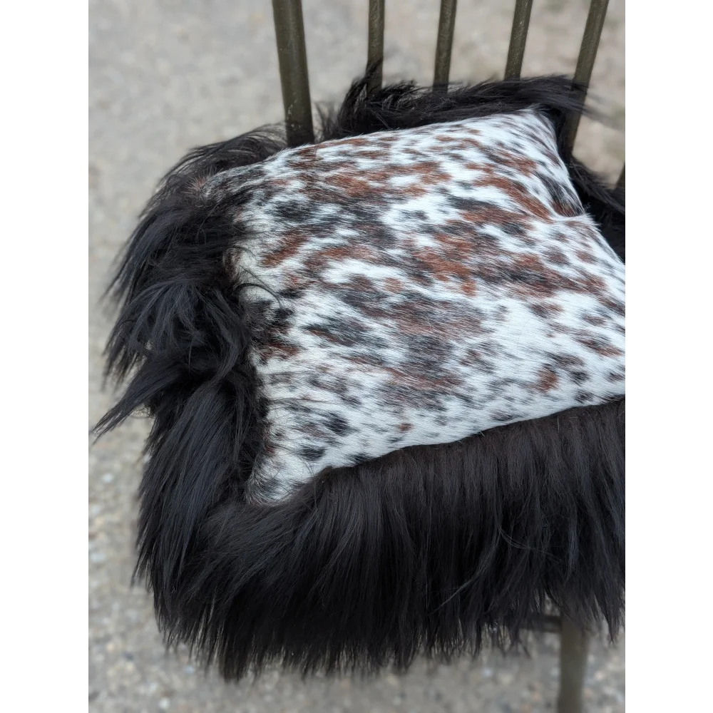 A plush square cushion in natural cowhide with white and brown speckles and long wool black icelandic trim