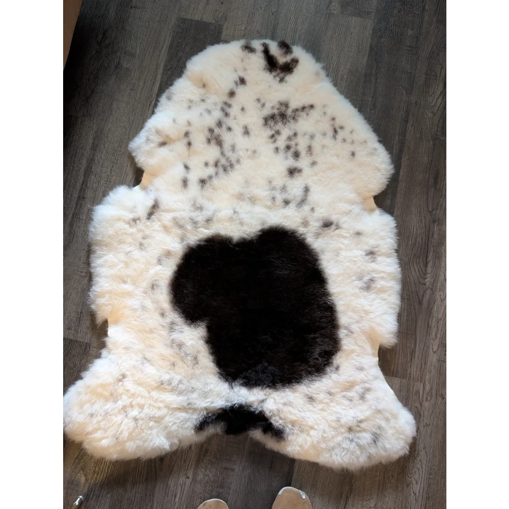 A plush cream-colored icelandic sheepskin rug with short wool featuring a black apple-shaped patch and dark speckles