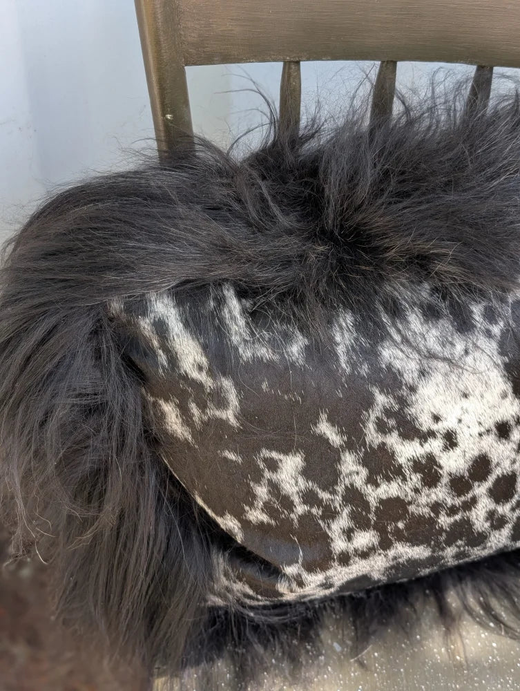 A plush black icelandic sheepskin throw with long wool draped over a cowhide pillow with white and dark brown spots