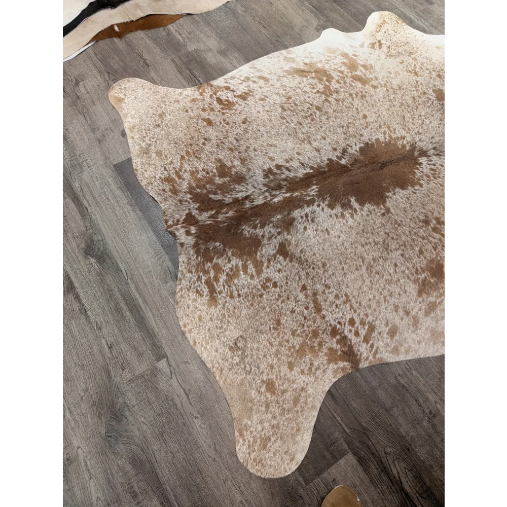 A large cowhide rug in natural tones with white and brown patches laid on a gray wood floor from angus oliver