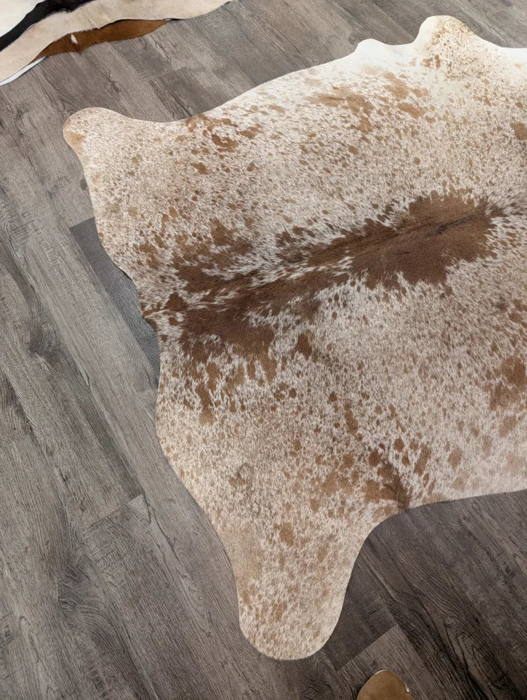 A large cowhide rug in natural tones with white and brown patches laid on a gray wood floor from angus oliver