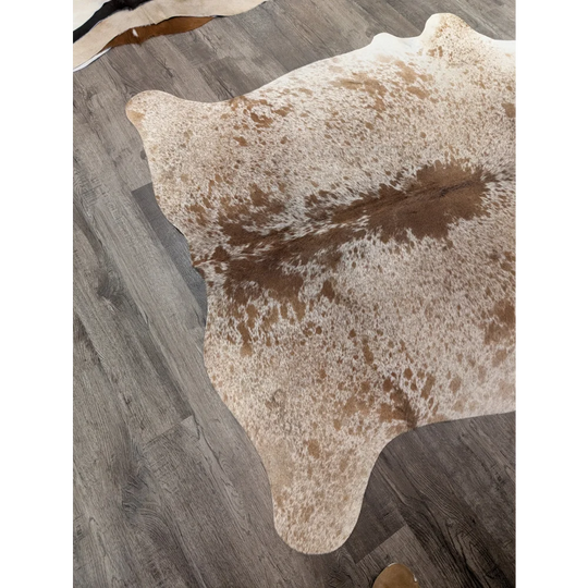 A large cowhide rug in natural tones with white and brown patches laid on a gray wood floor from angus oliver