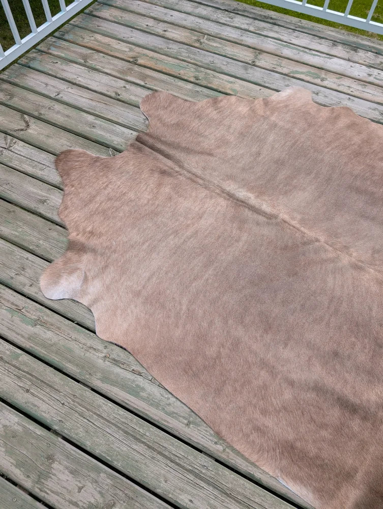 A large cowhide rug with unique characteristics in soft light brown natural fur lies flat on weathered wooden deck planks