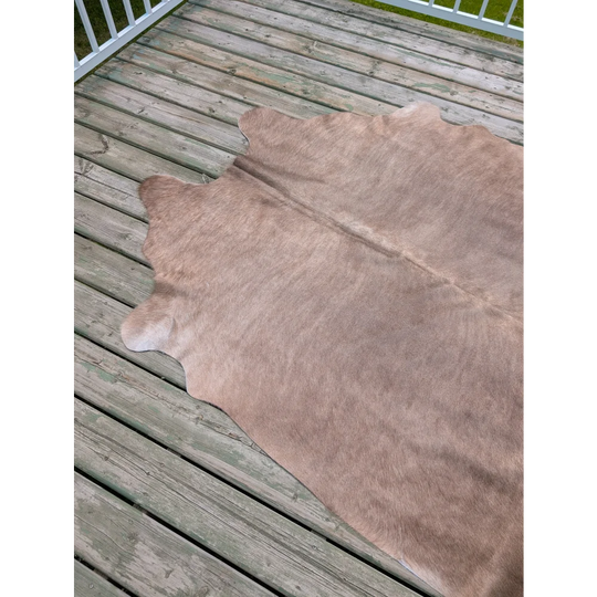 A large cowhide rug with unique characteristics in soft light brown natural fur lies flat on weathered wooden deck planks