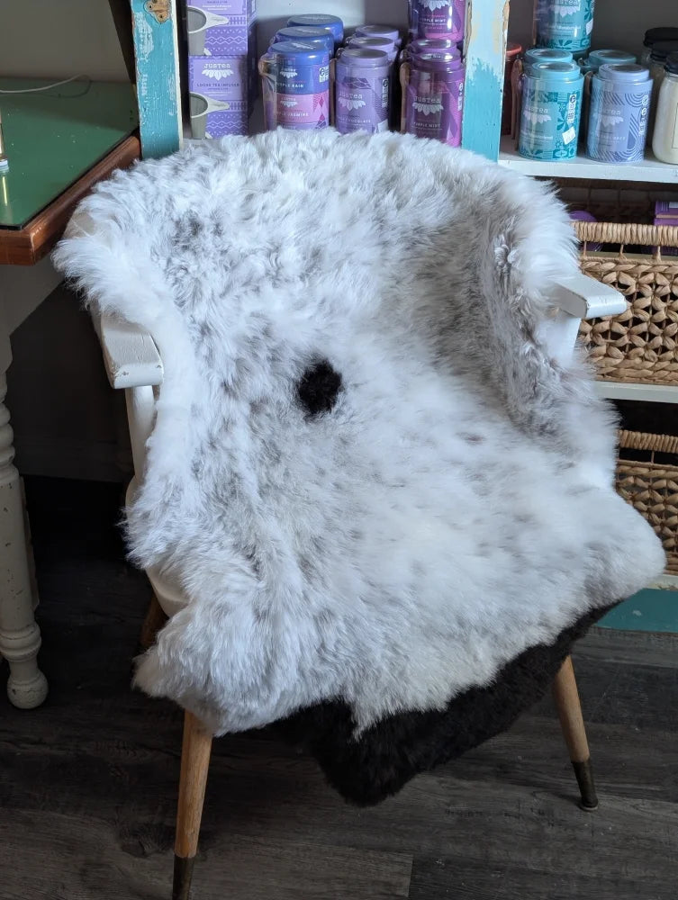 A fluffy white and gray medium spotted icelandic sheepskin rug draped over a wooden chair with a black spot