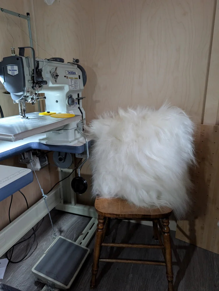The moo cushion is a white fluffy faux fur cushion with long wool texture resting on a wooden stool by angus oliver