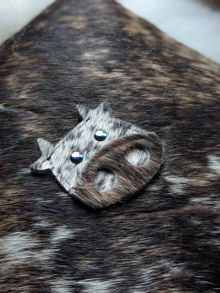 The moo cushion is a cow-shaped brooch made from gray and brown spotted leather with blue gemstone eyes inspired by angus oliver
