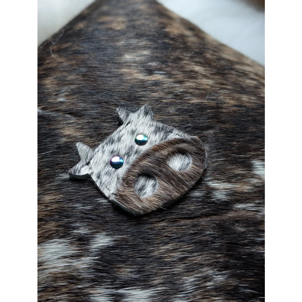 The moo cushion is a cow-shaped brooch made from gray and brown spotted leather with blue gemstone eyes inspired by angus oliver