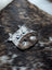 The moo cushion is a cow-shaped brooch made from gray and brown spotted leather with blue gemstone eyes inspired by angus oliver