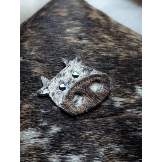The moo cushion is a cow-shaped brooch made from gray and brown spotted leather with blue gemstone eyes inspired by angus oliver