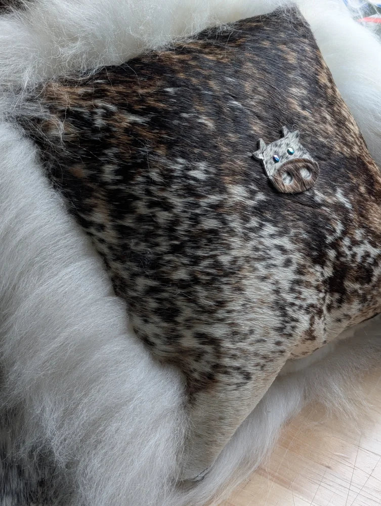 The moo cushion features a decorative cow-shaped brooch with blue eyes and a textured dark brown and white hide finish