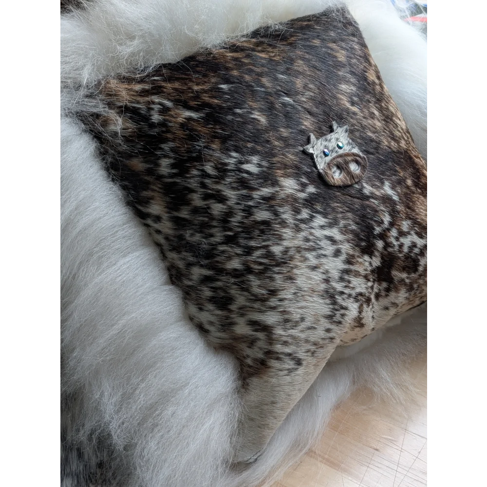 The moo cushion features a decorative cow-shaped brooch with blue eyes and a textured dark brown and white hide finish