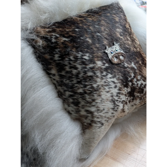 The moo cushion features a decorative cow-shaped brooch with blue eyes and a textured dark brown and white hide finish