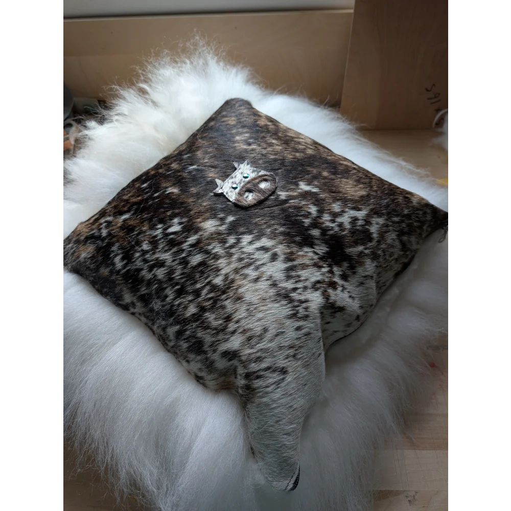 The moo cushion is a square cowhide cushion with natural brown and white spots and a silver cat figurine with green eyes