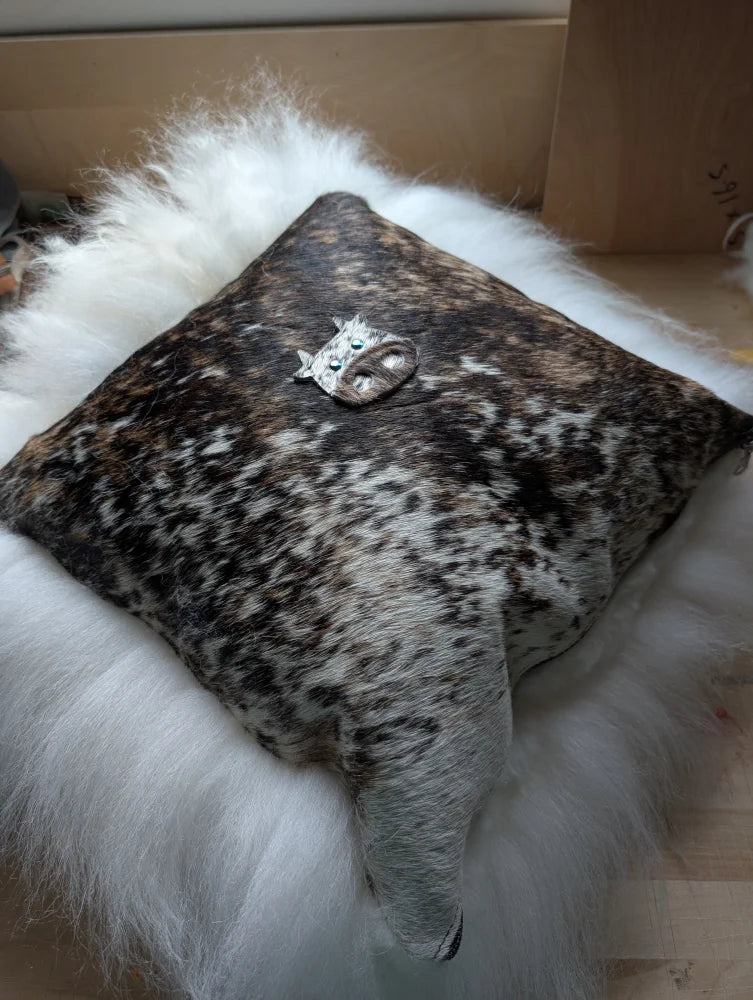 The moo cushion is a square cowhide cushion with natural brown and white spots and a silver cat figurine with green eyes