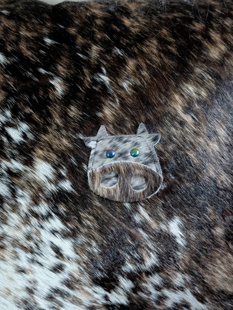 A cow-shaped brooch called the moo cushion made of brown and white spotted leather with blue and green gemstone eyes, perfect for angus oliver fans
