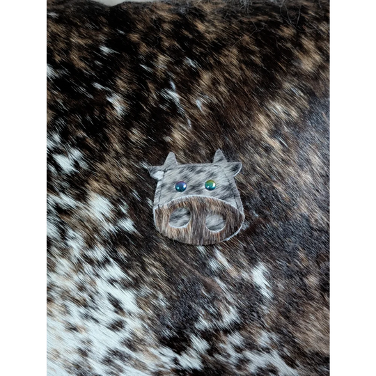 A cow-shaped brooch called the moo cushion made of brown and white spotted leather with blue and green gemstone eyes, perfect for angus oliver fans