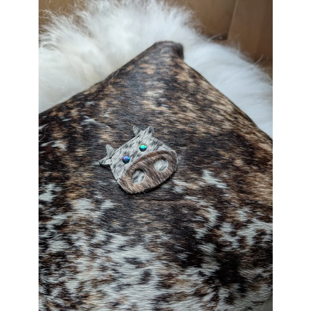 The moo cushion is a small rustic cow-shaped brooch made of brown and white spotted leather with iridescent blue-green eyes, perfect for angus oliver fans and those who love sewing machines and long wool style