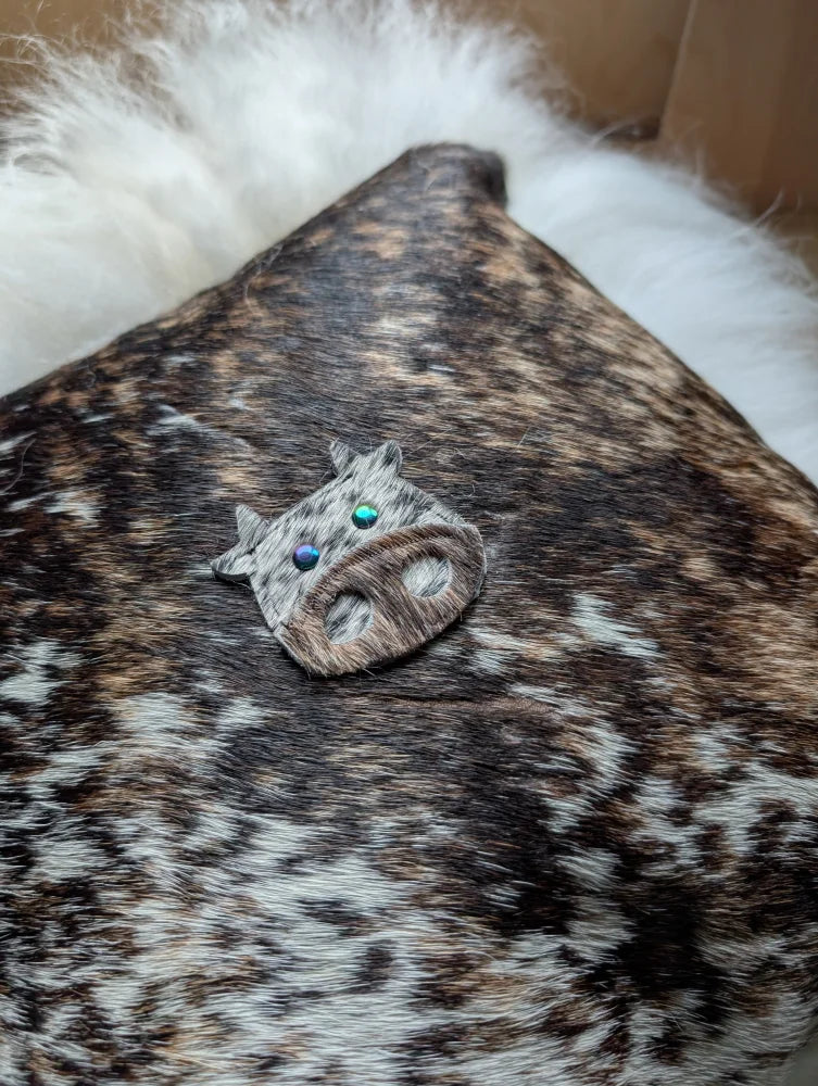The moo cushion is a small rustic cow-shaped brooch made of brown and white spotted leather with iridescent blue-green eyes, perfect for angus oliver fans and those who love sewing machines and long wool style