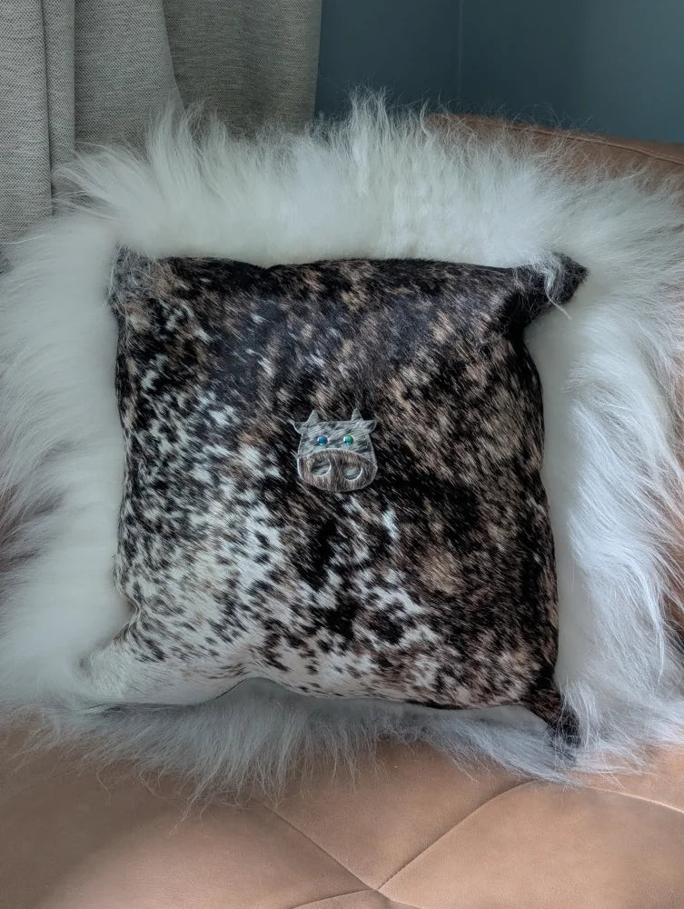 The moo cushion is a square black and white speckled cowhide pillow with plush white faux fur trim and a silver cat brooch