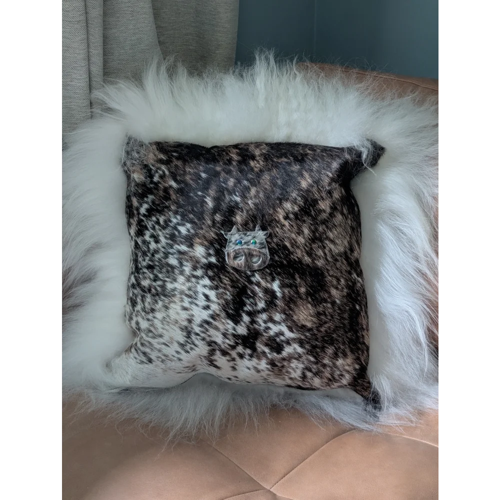 The moo cushion is a square black and white speckled cowhide pillow with plush white faux fur trim and a silver cat brooch